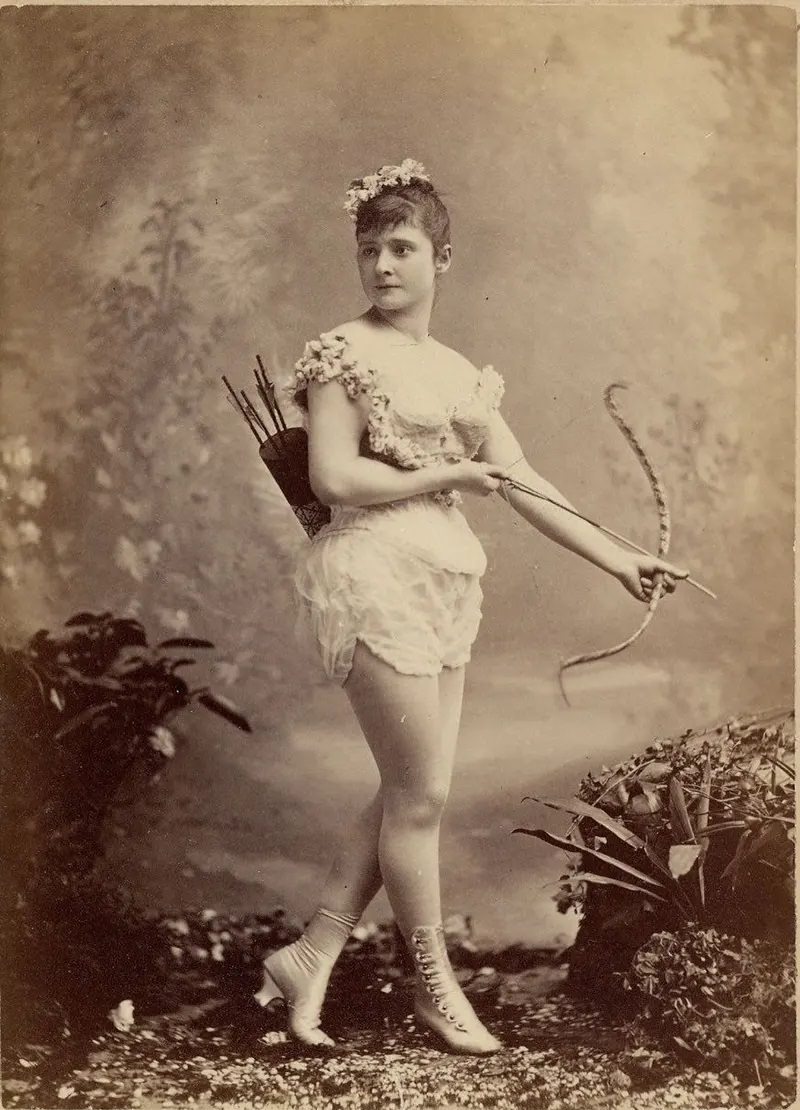 #21 Miss Farrington in a short sleeveless costume, holding bow and arrow, with quiver and arrows on back, buttoned and heeled boots.
