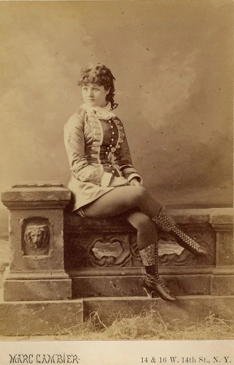 #6 Minnie Marshall sitting on a stone fence, in a short dress and boots.