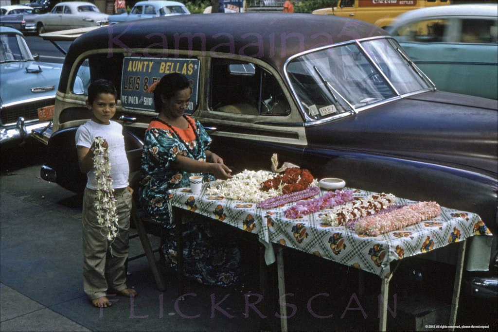 #70 Aunty Bella’s sidewalk lei stand in its usual spot on Kalakaua Avenue across from the Waikiki Theatre, 1957