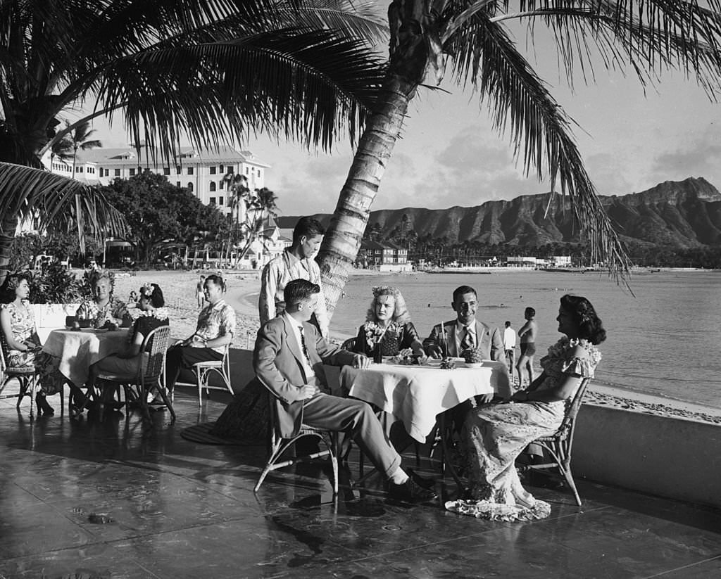 #59 Couples sit outdoors at patio tables, having a meal at a beach side restaurant while a waiter looks on, Waikiki Beach, 1945
