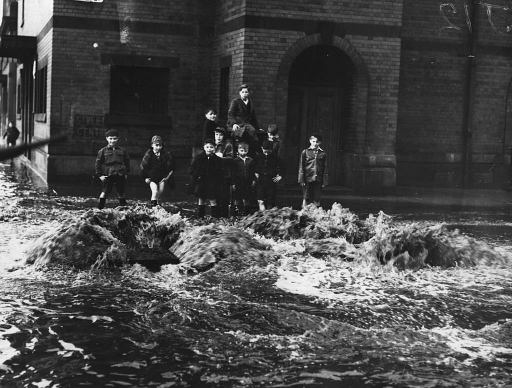 #73 Children watch as a torrent of water pours out of drains in a Glasgow street during severe floods throughout Scotland, 1964