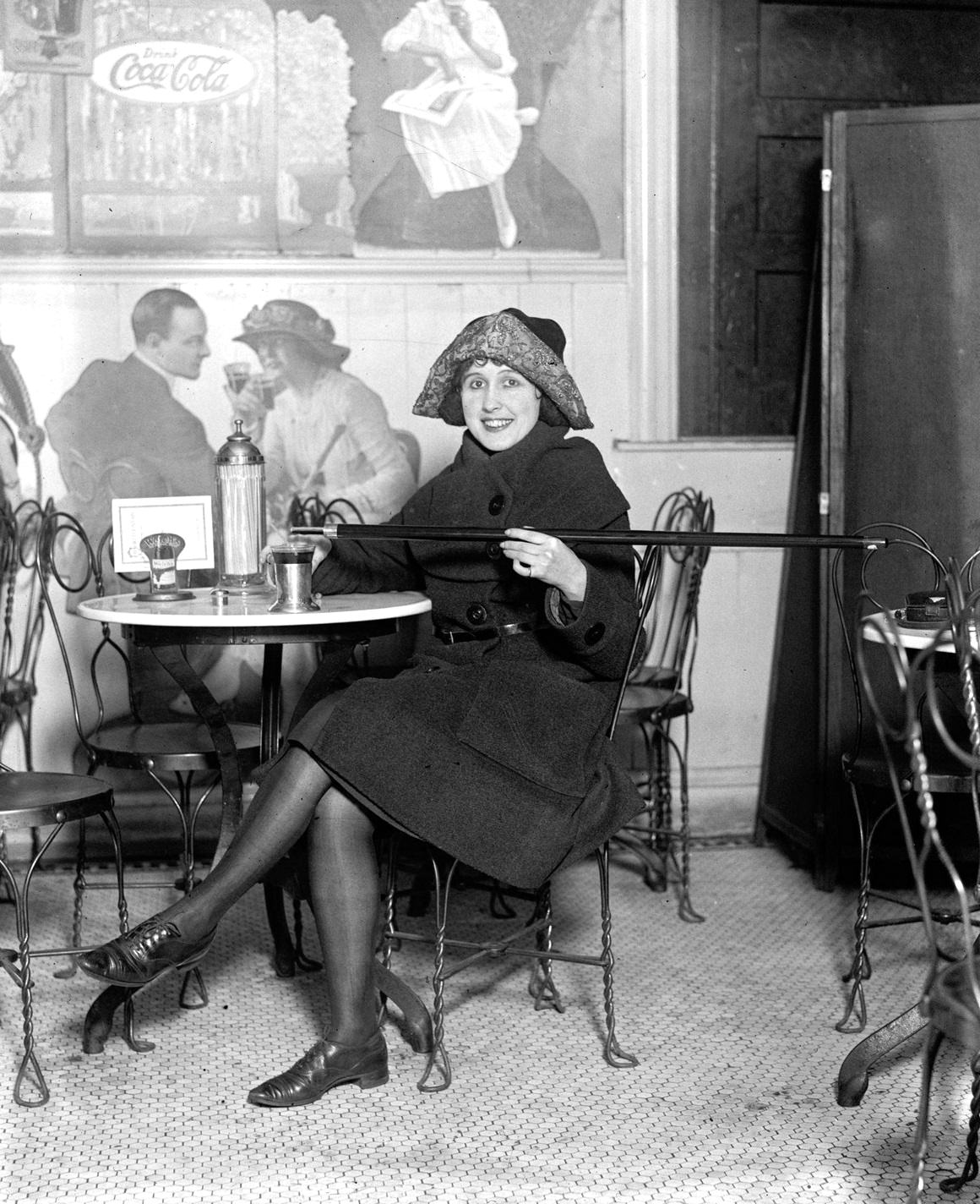 #6 A woman in Cloche Hat, seated at a soda fountain table, as she pours alcohol from a cane into a cup during Prohibition, 1922