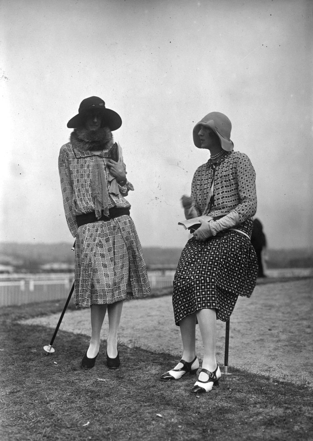 #114 Two racegoers wearing long-waisted patterned dresses with panels One has two-tone one-bar shoes and her cloche hat has a wide turned-down brim at left-hand side, 1925