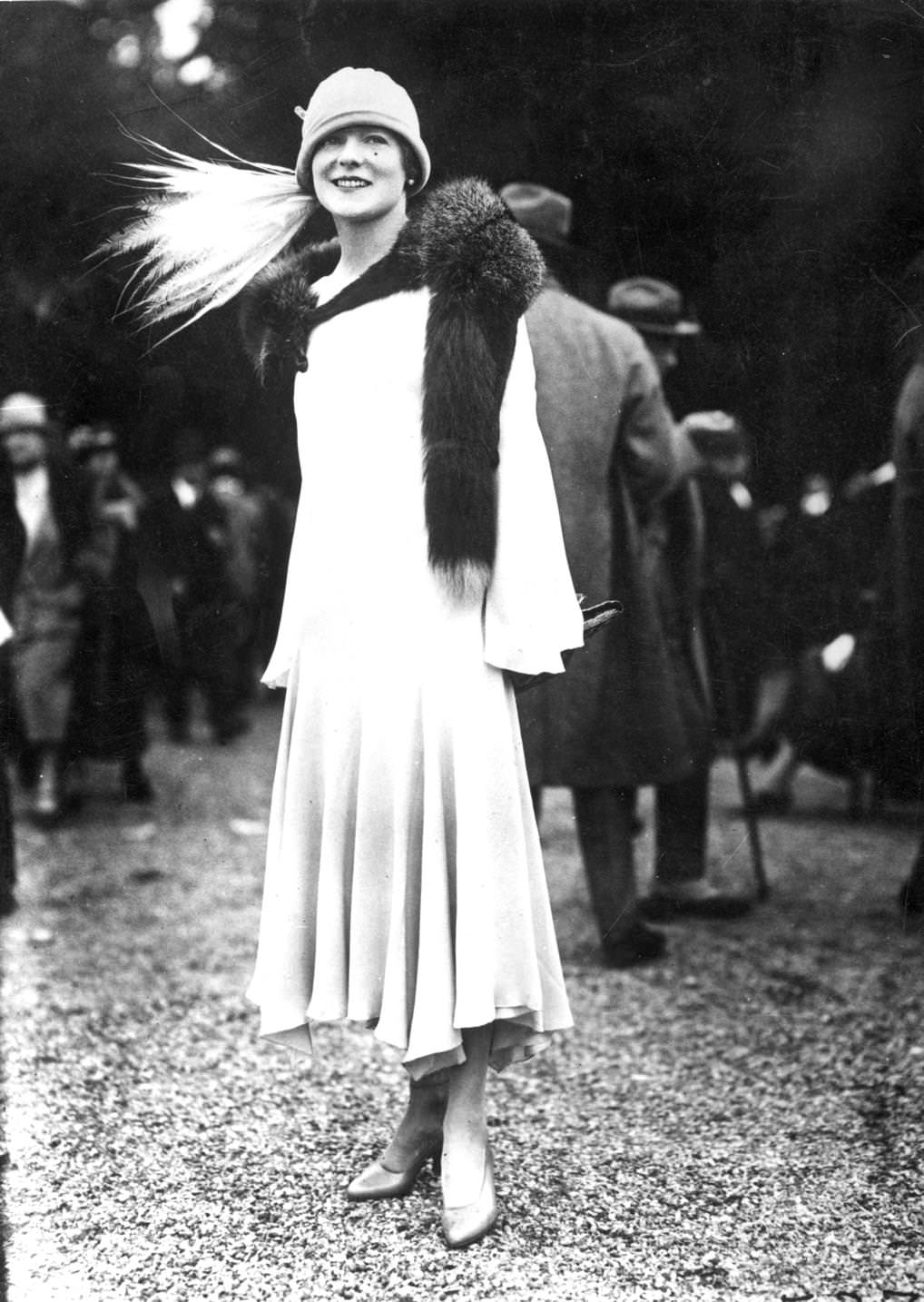 #123 An elegant evening dress with a tapering uneven hemline and sleeves. A fur stole and hat with an array of long white feathers complete the look, 1925