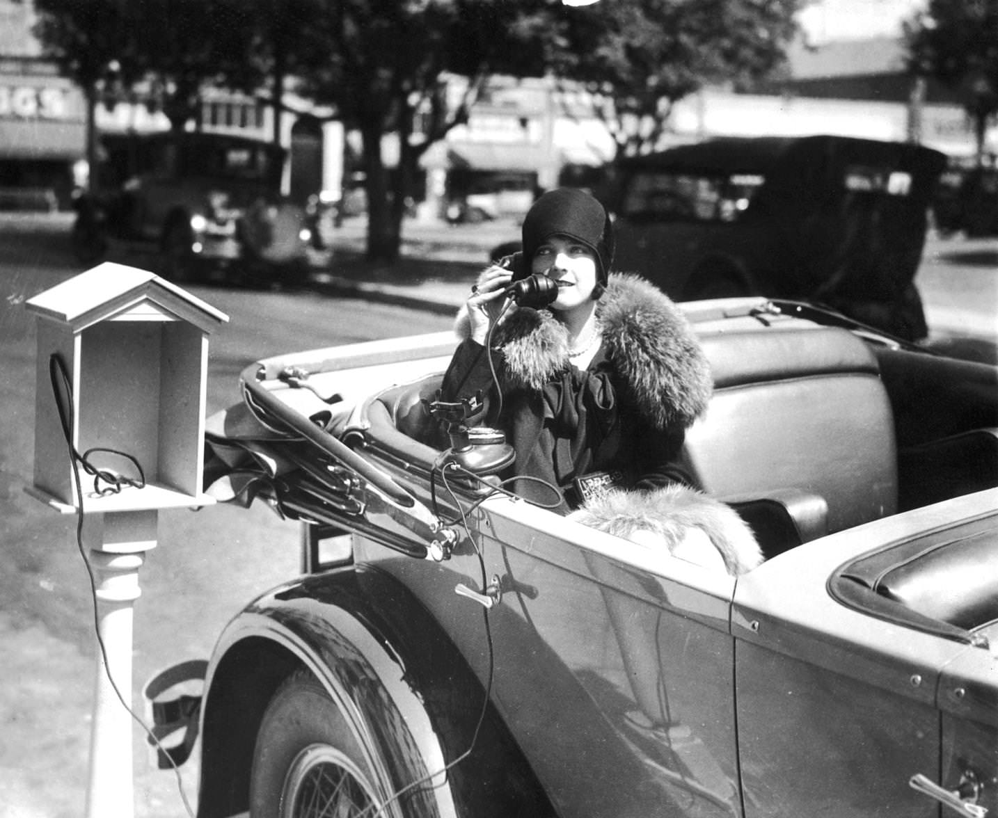 #133 A woman sits in an automobile while talking on the telephone from a phone service stand at the Hotel Constance in Pasadena, California. She is wearing a cloche hat and a fur-trimmed coat, 1925
