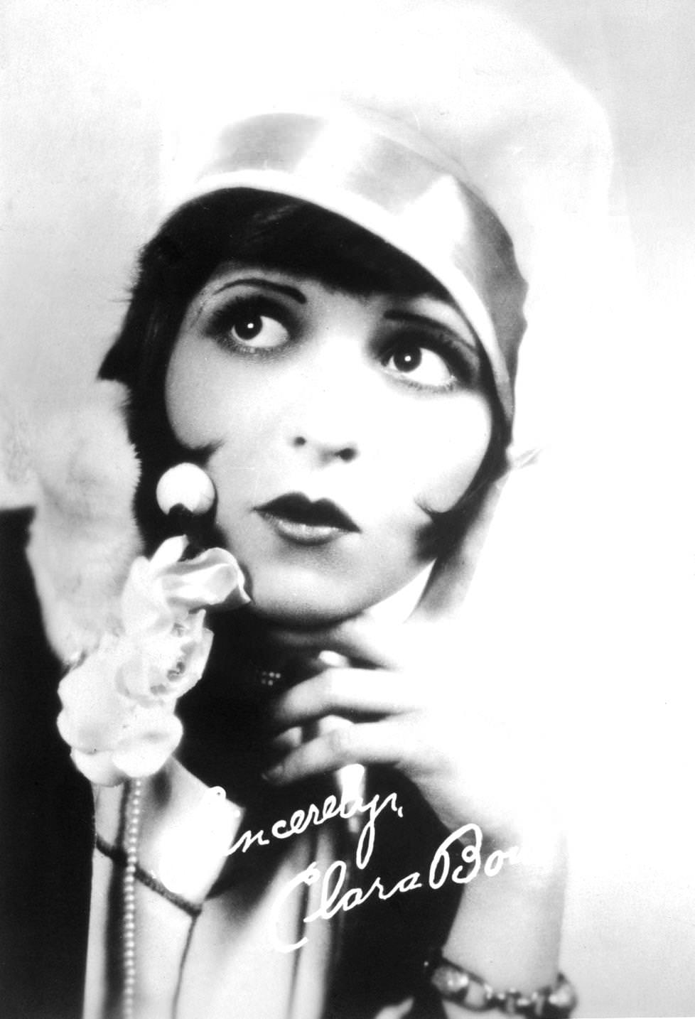 #134 Autographed studio portrait of American actor Clara Bow wearing a cloche hat and a bobbed hairstyle while holding her hand to her face and pursing her lips, 1925