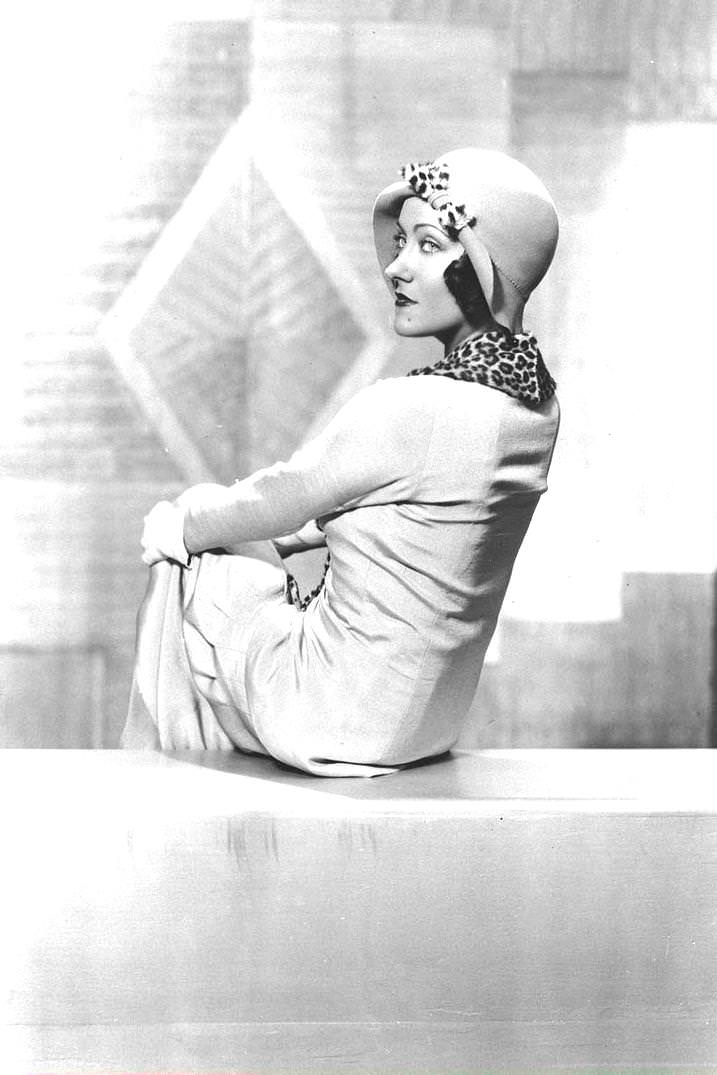 #135 American film actor and producer Gloria Swanson sits with her knees crossed and her back to the camera as she looks over her shoulder. She wears white gloves, a cloche hat with a leopard print bow, and a matching scarf, 1926