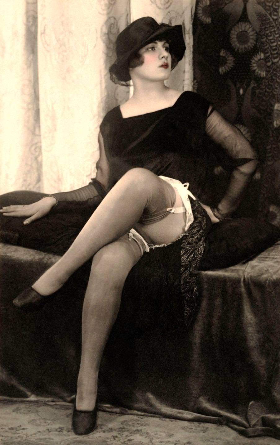 #18 A woman seated on a divan, she wears a square necked black velvet dress with sheer chiffon sleeves and a black cloche hat, 1920
