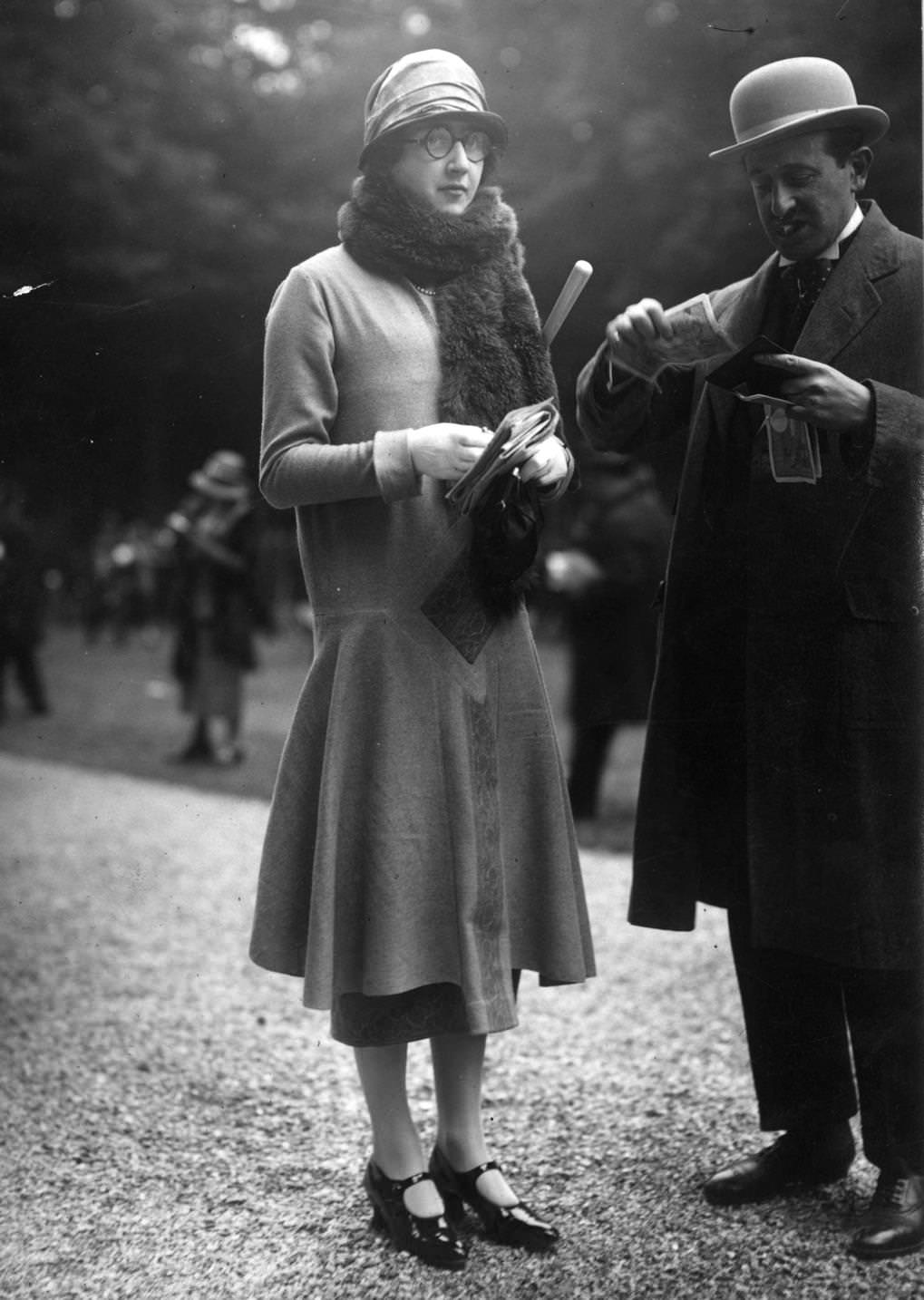 #146 A woman wears a plain long waisted seven eighths coat worn with a cloche hat and a fur scarf. Her male friend puts away some large banknotes, 1924