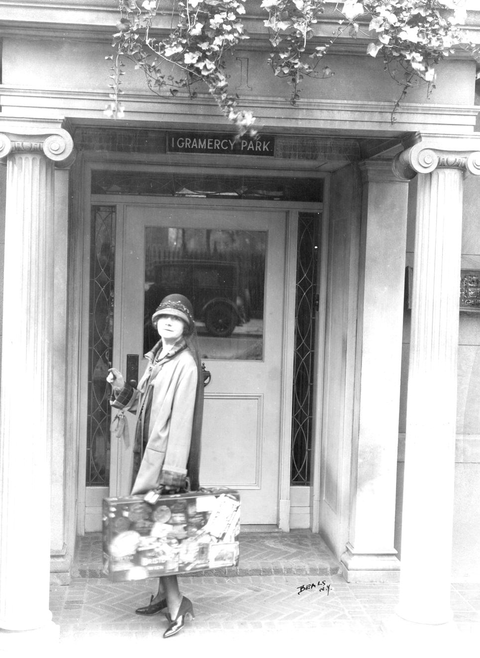 #7 Journalist Mabel Herbert Urner, in cloche hat, standing on pillared stoop of her 1 Gramercy Park house, holding suitcase covered with travel stickers, New York, 1922.