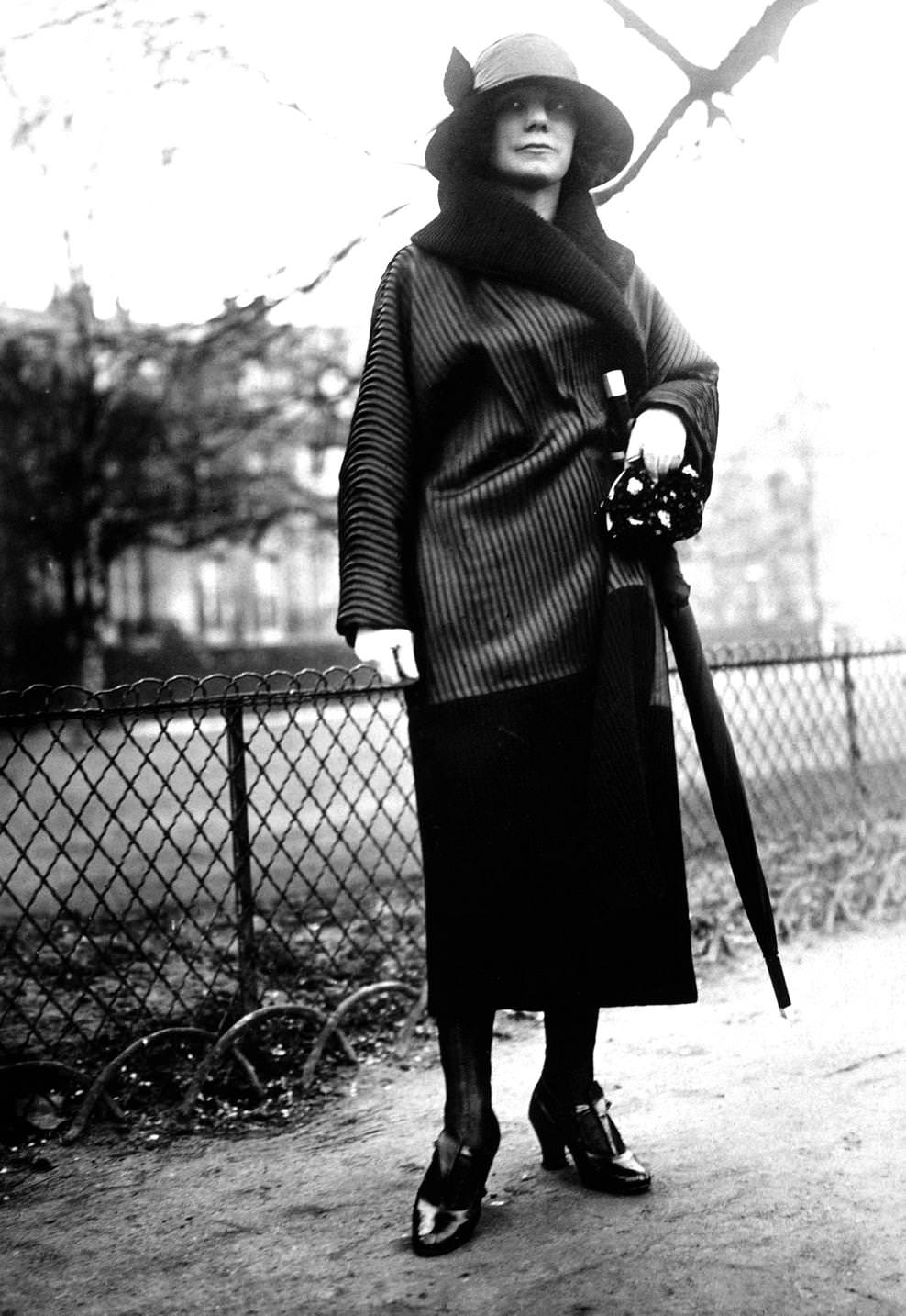 #9 Fashion model wearing a short wrapover coat with fur collar, brimmed cloche hat, straight skirt and carries an umbrella, 1920s