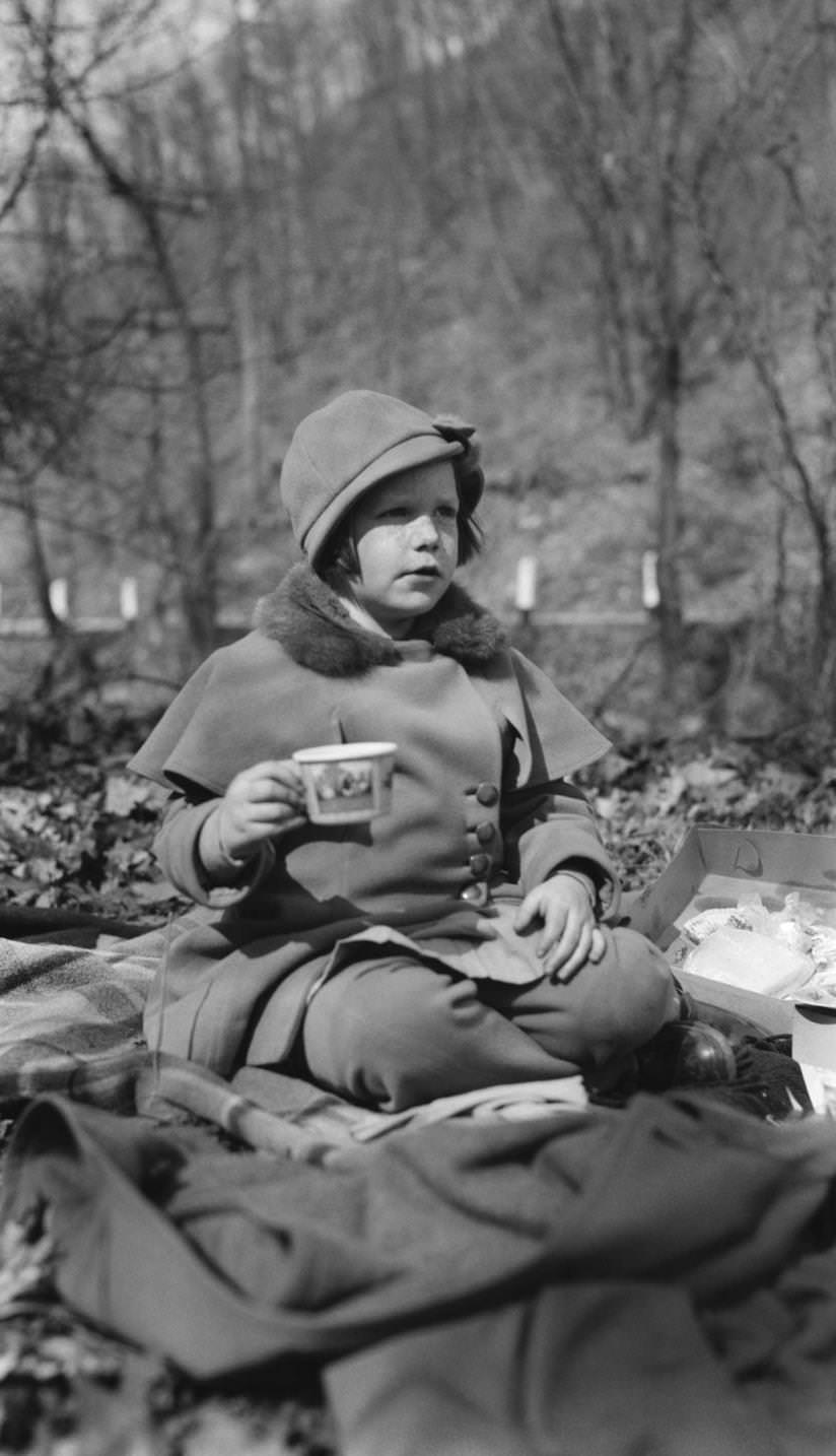 #55 Young, smartly dressed child (girl), wearing caped coat and cloche type hat, sitting on blankets outdoors having a picnic, 1924.