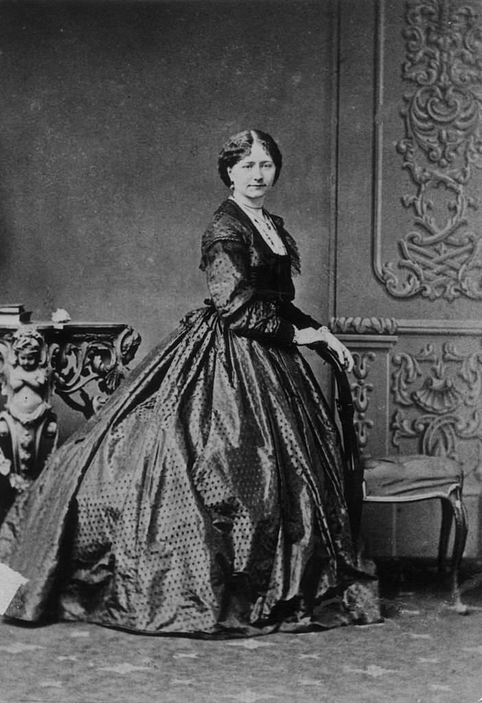 #21 A young woman waits patiently whilst the hoops to support her crinoline are prepared, 1860
