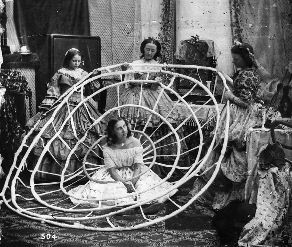 #22 A woman wearing a crinoline being dressed with the aid of long poles to lift her dress over the hoops.