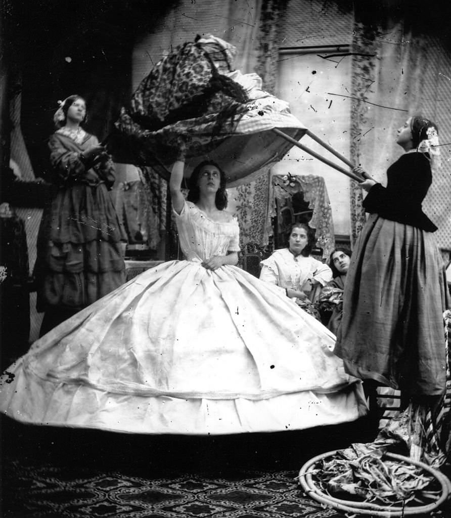 #23 A woman wearing a crinoline being dressed with the aid of long poles to lift her dress over the hoops.