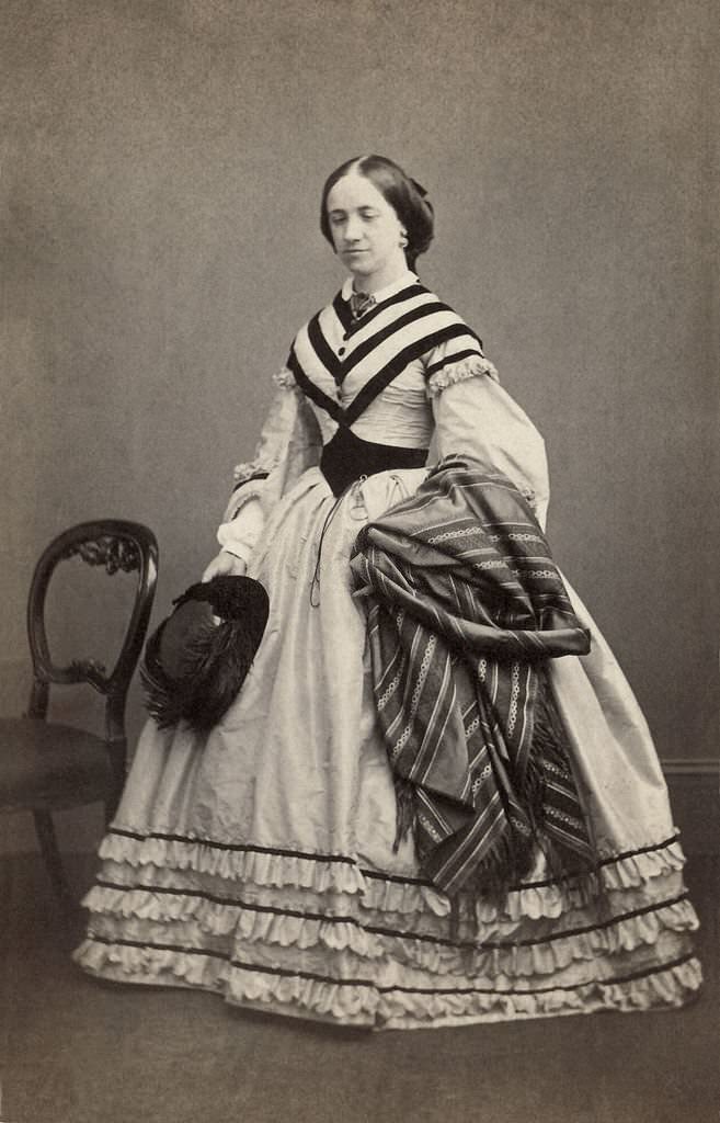 #37 The woman carries a feathered hat and a piece of striped fabric, possibly a shawl. She wears a monocle or pince-nez at her waist, 1856