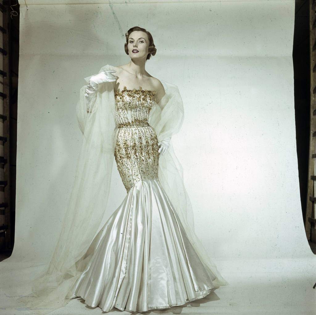 #97 A model presenting for Jacques Fath, a gold-embroidered satin sheath dress, which flares out in godets at the bottom.