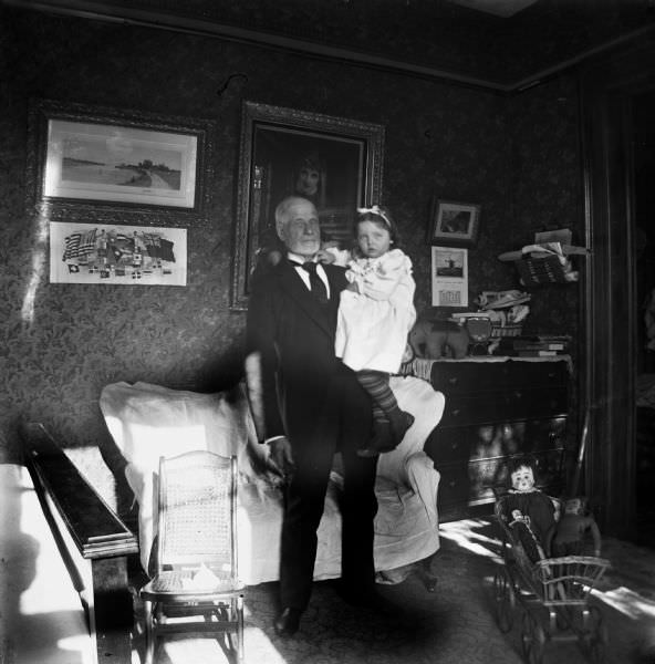 #51 An elderly man stands in a room holding a small girl, 1898