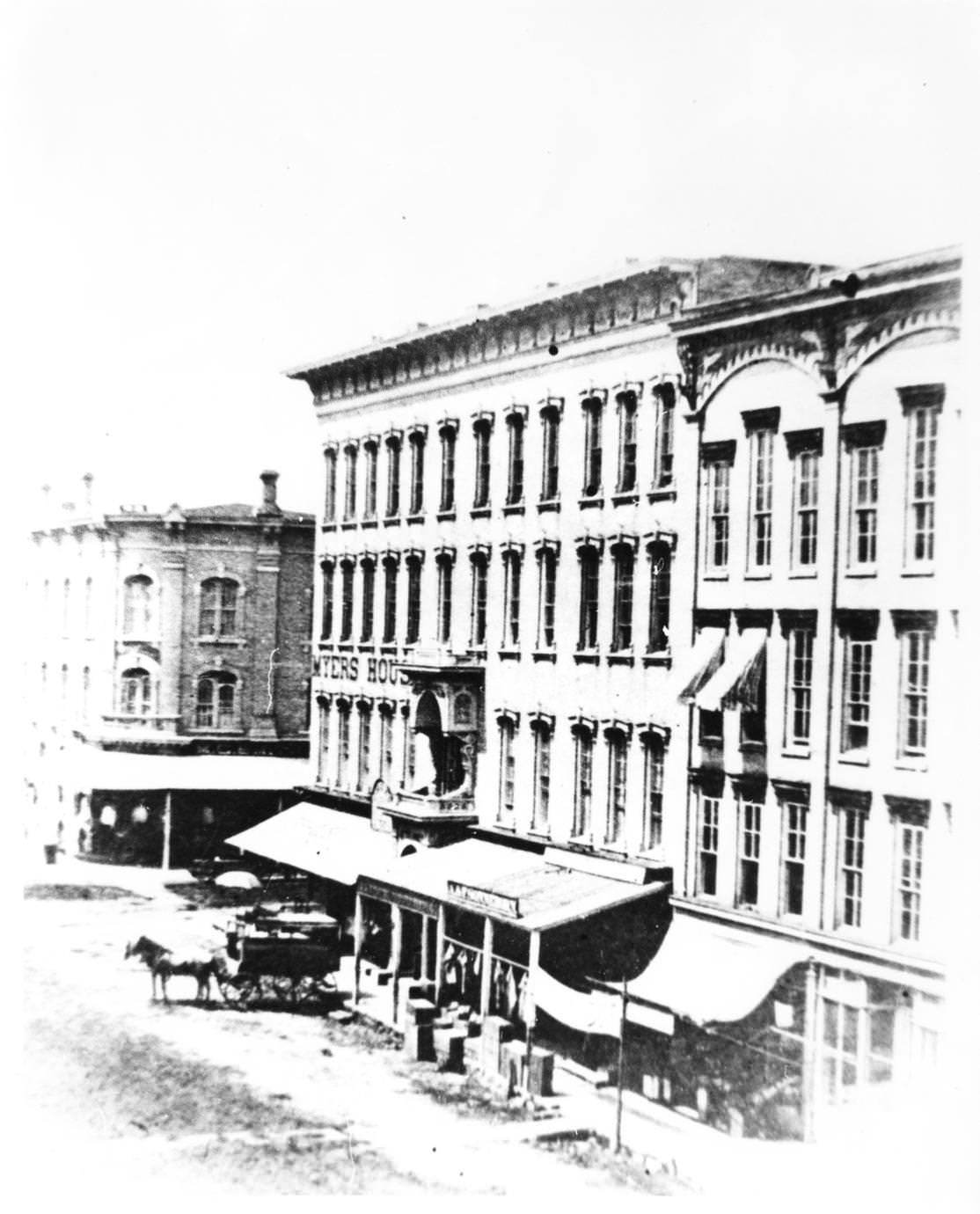 #65 Myers House Hotel from South Main Street. Photo includes an unpaved street, a horse and coach, 1875