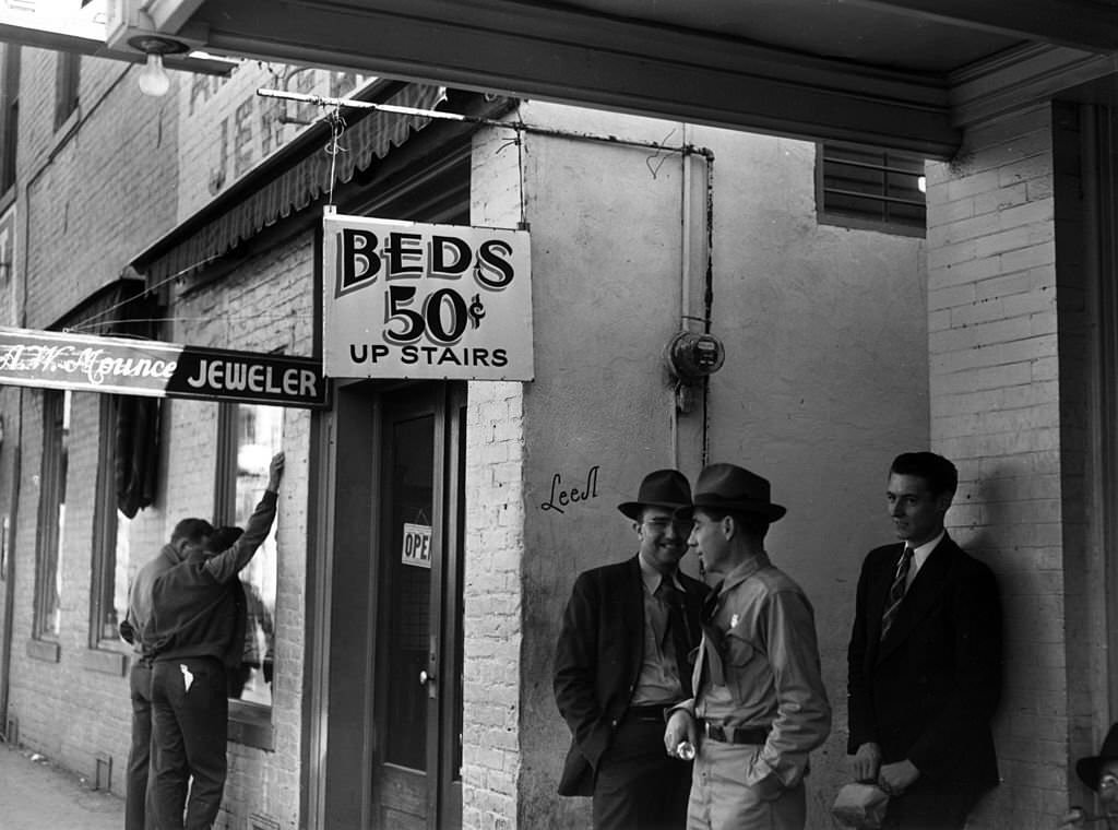 #121 Workers standing outside of a housing facility where the sign reads ‘Beds 50 cents up stairs’, December 1940