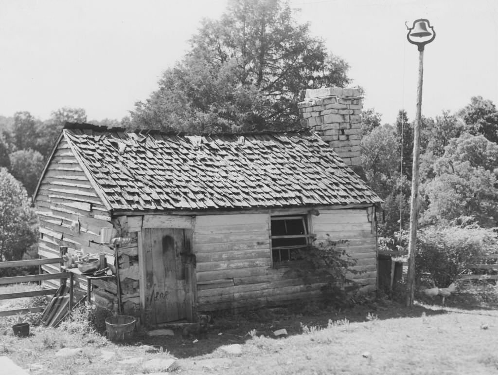#222 A small wooden shack, with a shingled roof, brick chimney stack, and a bell set on top of a tall pole at right, Kentucky, 1940