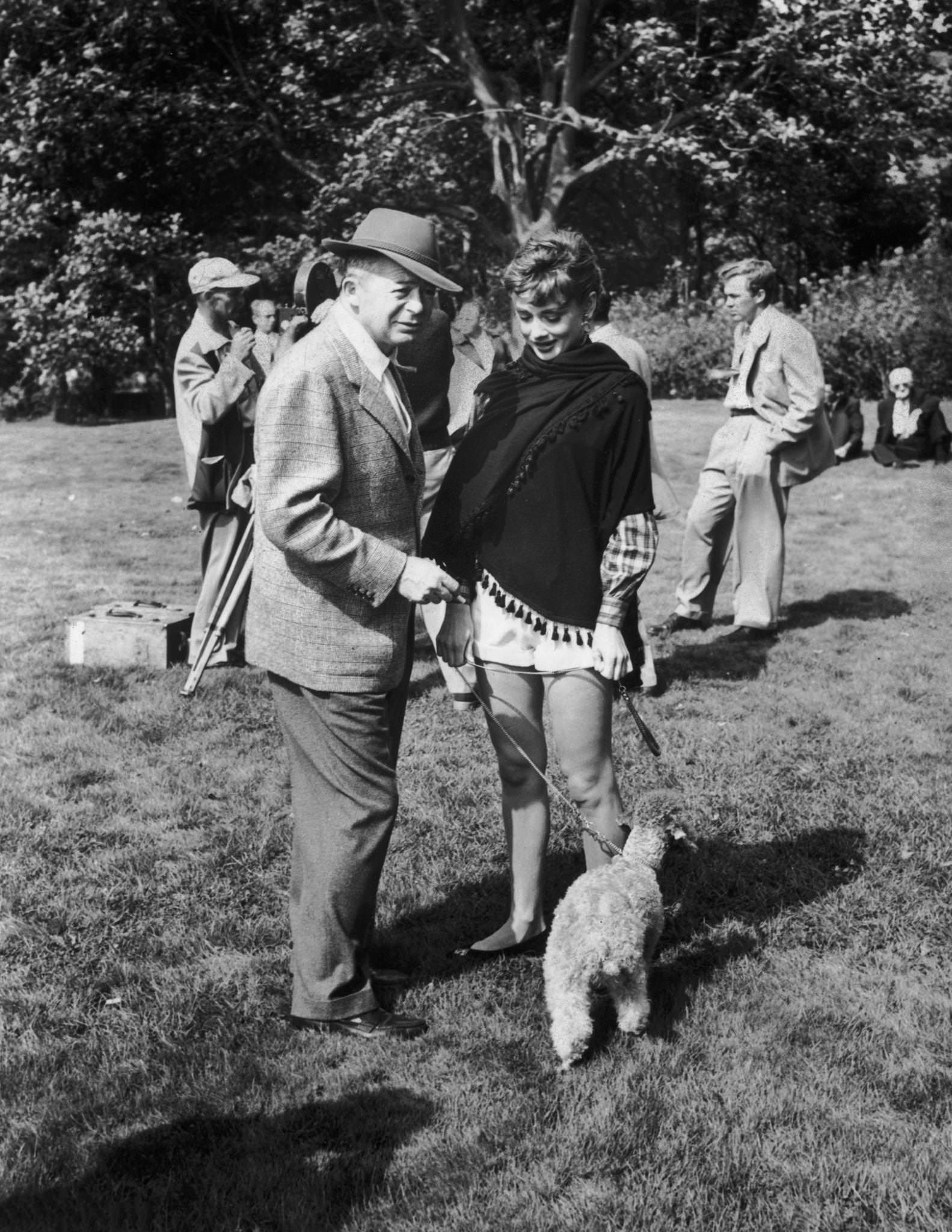 Director Billy Wilder standing outdoors beside Belgian-born actor Audrey Hepburn, who holds a poodle on a leash, on location for his film, ‘Love in the Afternoon’.