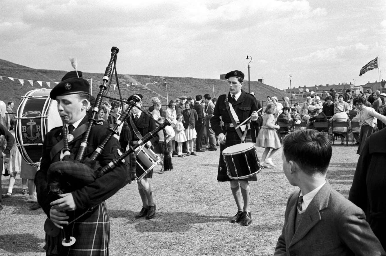 #92 A village festival, with the band; in a close-up, there is a bagpipe player, who wears the kilt, and on the background is a crowd, Scotland, 1968