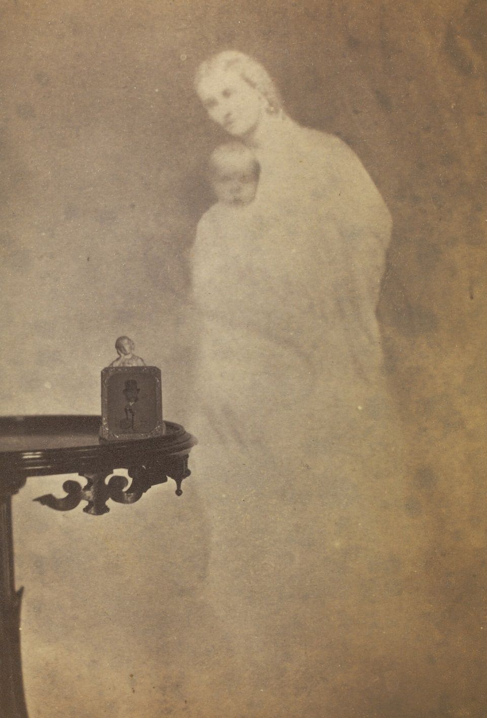 #2 Photograph of a statuette and drawing on a side table, with a faint image of a woman and baby in the background.