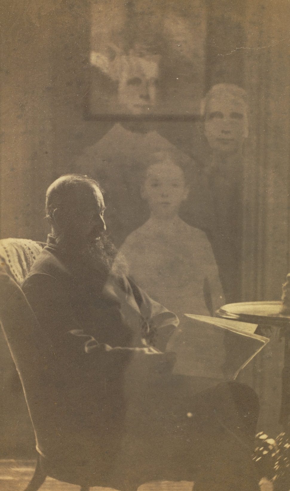 #22 Profile portrait of a man with a beard seated in an upholstered chair, next to a side table. The faint images of three children appear behind him.