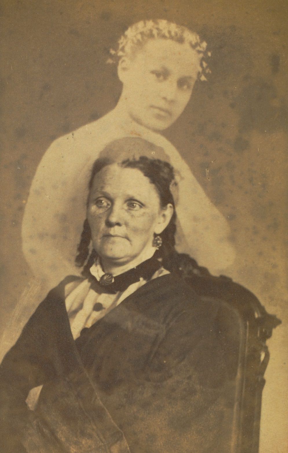 #23 Portrait of a woman seated with her arms crossed. The faint image of a younger woman wearing a floral hair wreath appears above her.
