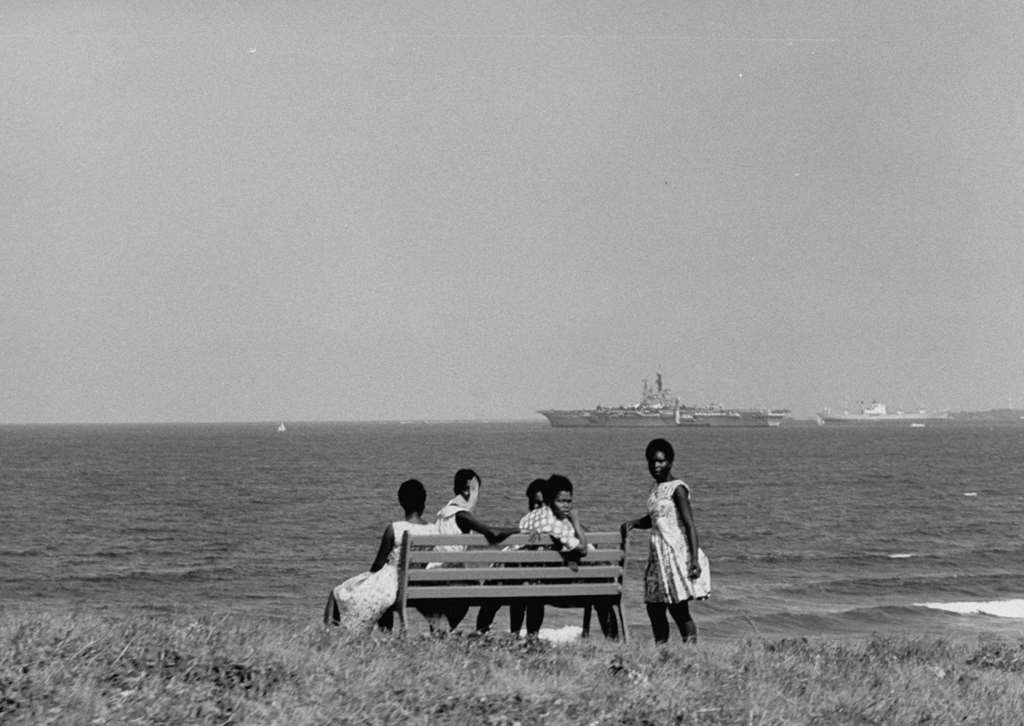 #24 Local residents seated on a bench to watch the Royal Navy light fleet carrier HMS Centaur (R06) on patrol off the coast of Dar es Salaam, 1964