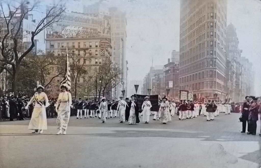 #34 Women wearing white Edwardian clothing, and sashes, and holding a US flag and pennants, followed by a marching band, taking part in a suffrage parade, 1913
