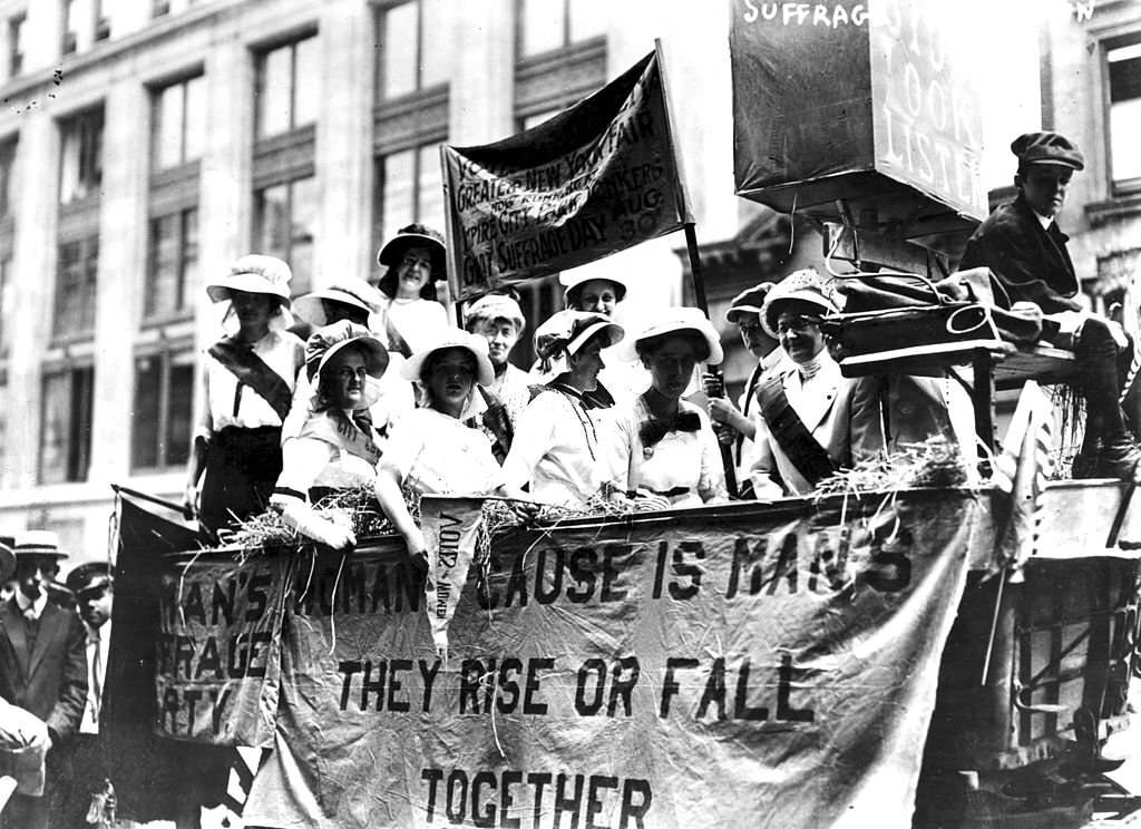 #49 Women riding in a Hay Wagon float in a suffrage parade, 1913. A banner hanging upon the float quotes Tennyson’s phrase ‘The Woman’s Cause Is Man’s; They Rise Or Fall Together’.