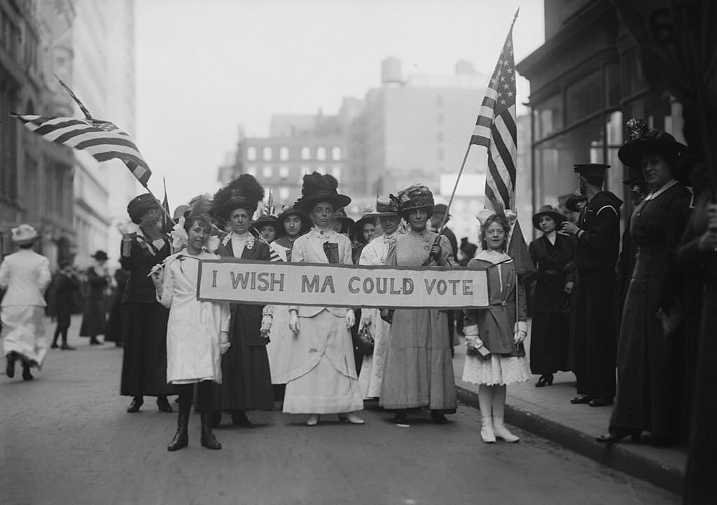 #56 A group of Women’s Suffrage activists march in a parade carrying a banner reading ‘I Wish Ma Could Vote’ circa 1913.