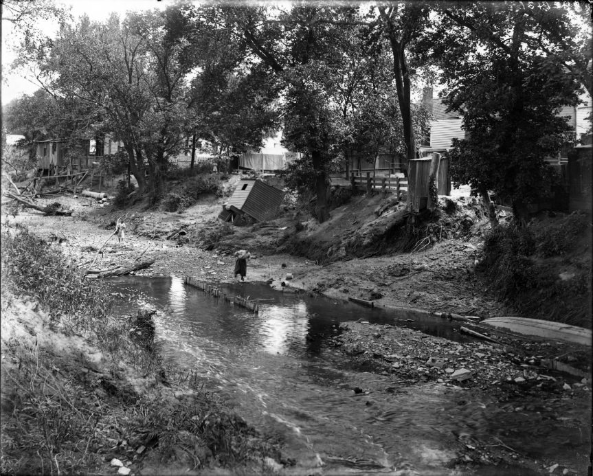 #131 River at low level running through a neighborhood, 1906. People are in the riverbed and a shack that has fallen off its foundation is also visible along the bank.