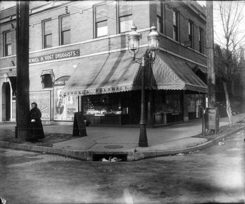 #132 Mengel & Vogt pharmacy at Taylor and Page Avenues, 1906. A woman is standing next to a pole in front of the building. The building still exists and appears to be vacant.