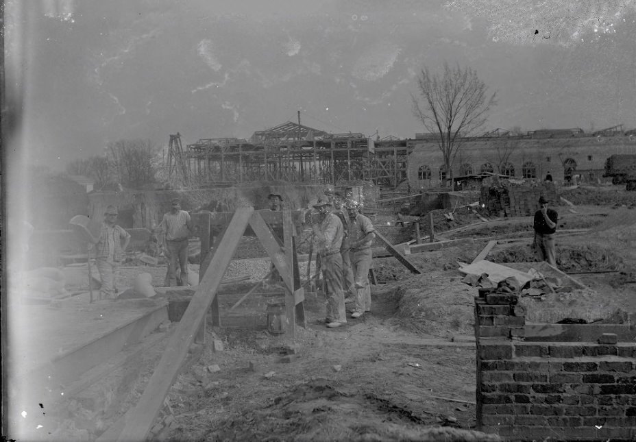 #238 A busy construction site shows the wide skeleton of a building in the background, stacks of bricks and lumber are in the middle ground, 1907