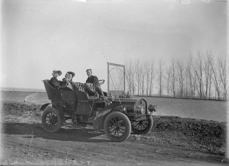 #250 Three people in a company care parked by the river, 1909