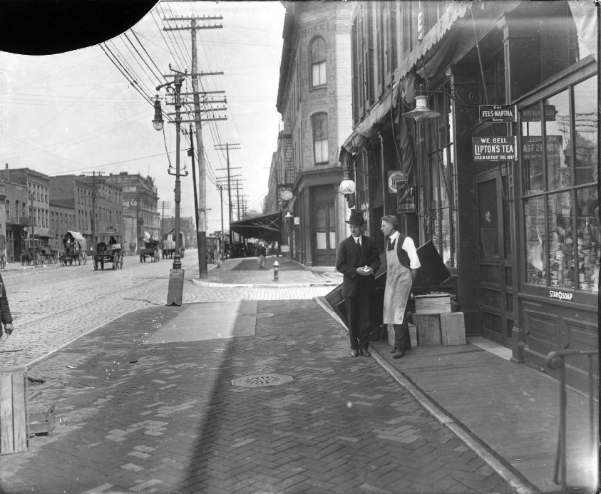 #90 Two men standing on the sidewalk of a major street. One man appears to work at the grocery they are standing in front of, 1904