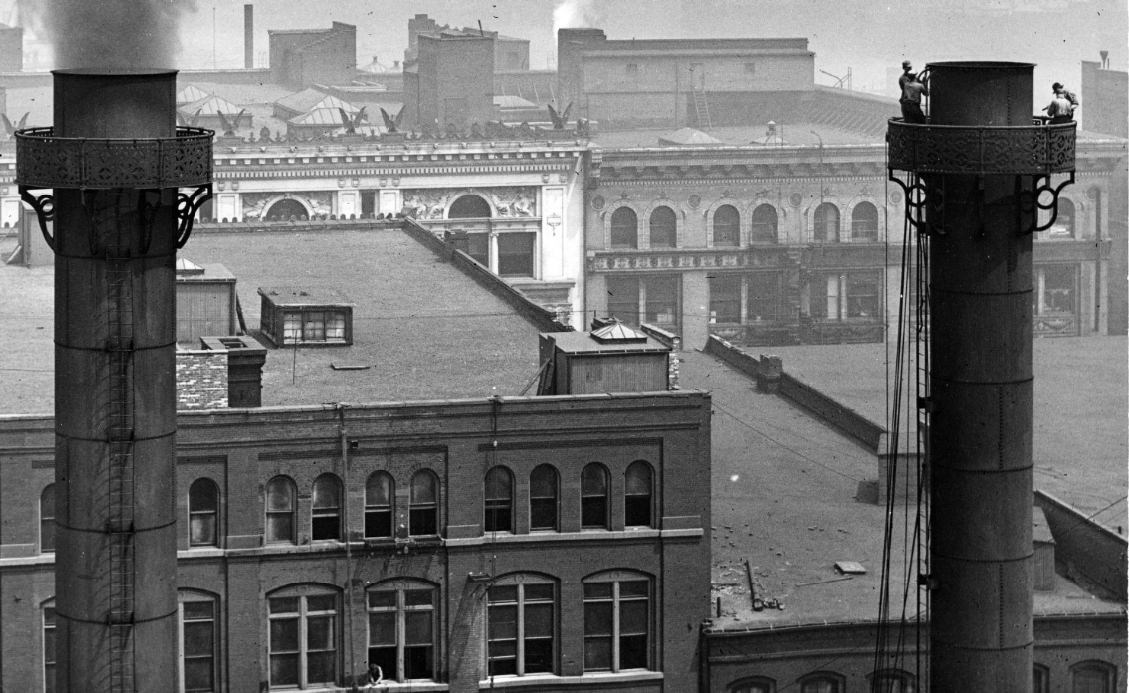#100 A pair of smokestacks, one of which is being worked on by a group of men near the top of the stack, 1908