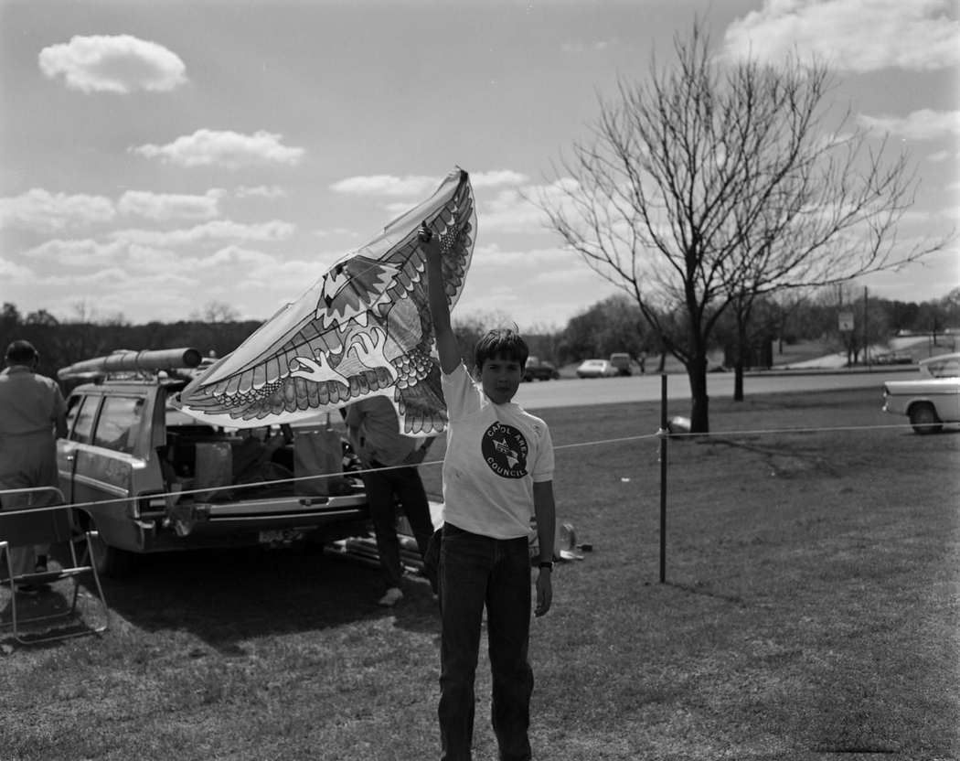 #65 Zilker Park Kite Tournament,March 8, 1970. A child is seen holding up a kite and a vehicle is parked behind him.