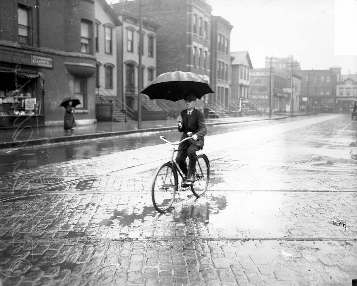 #97 Man holding an umbrella and riding a bicycle in the rain on an empty street, with visible streetcar tracks, during a streetcar strike, Chicago, Illinois, June 15, 1915.