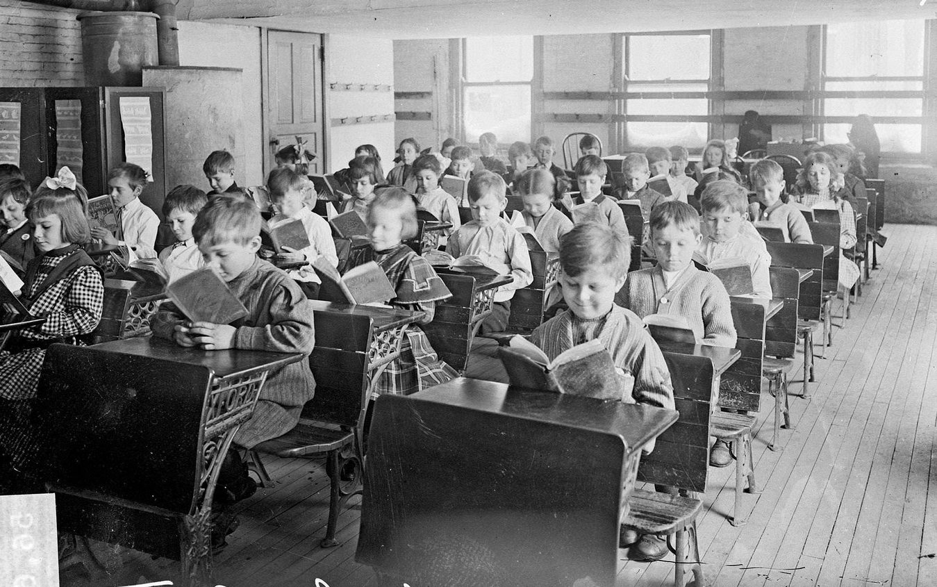 #133 Children reading at desks in a classroom at the Robert Emmet School, looking at the children from the front of the classroom in the Austin community area, Chicago, Illinois, 1910s.