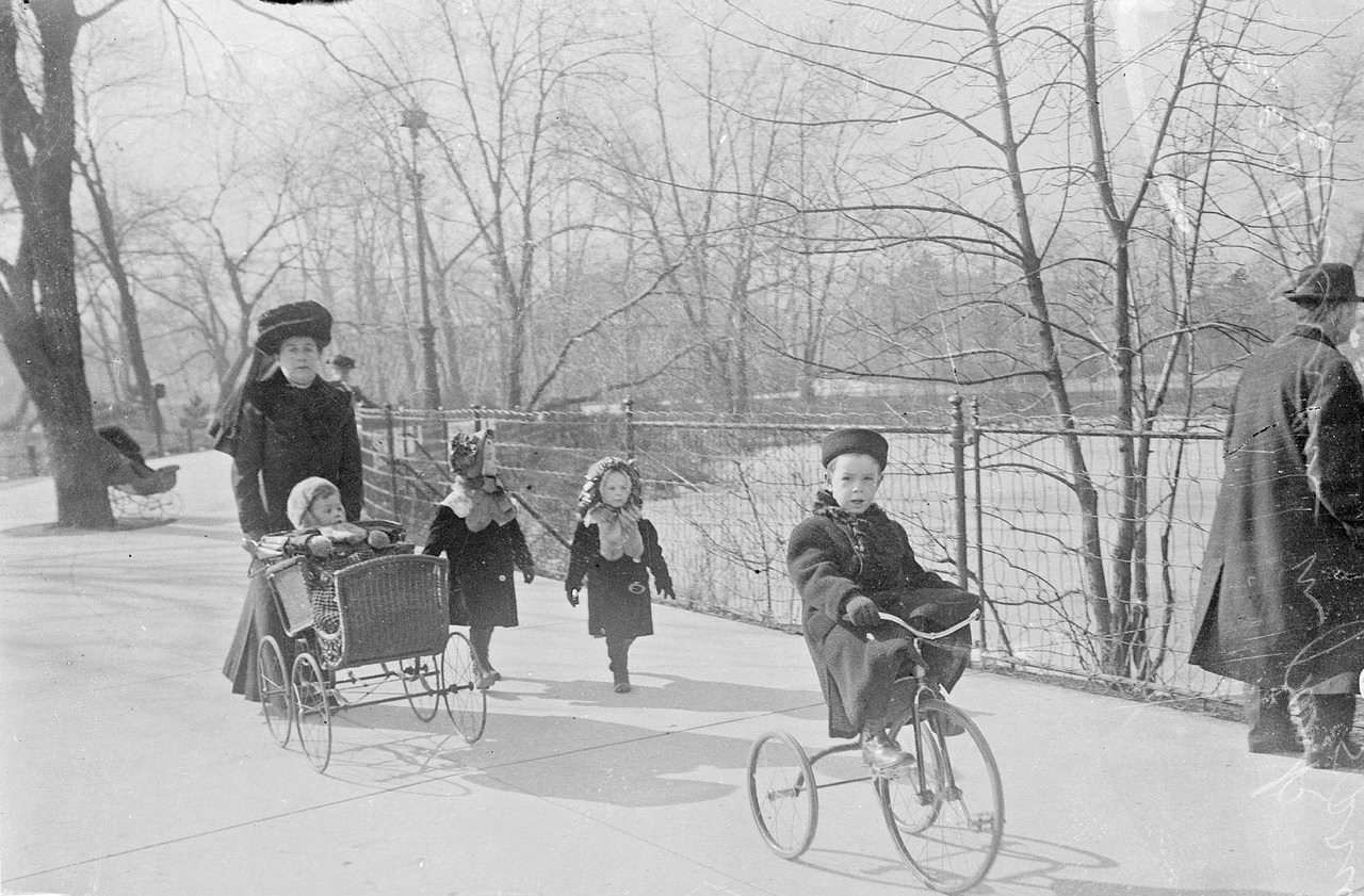 #135 A woman pushing a child in a stroller, with two children walking to the side and a child riding a tricycle in front of them in Lincoln Park in spring, Chicago, Illinois, 1910s.