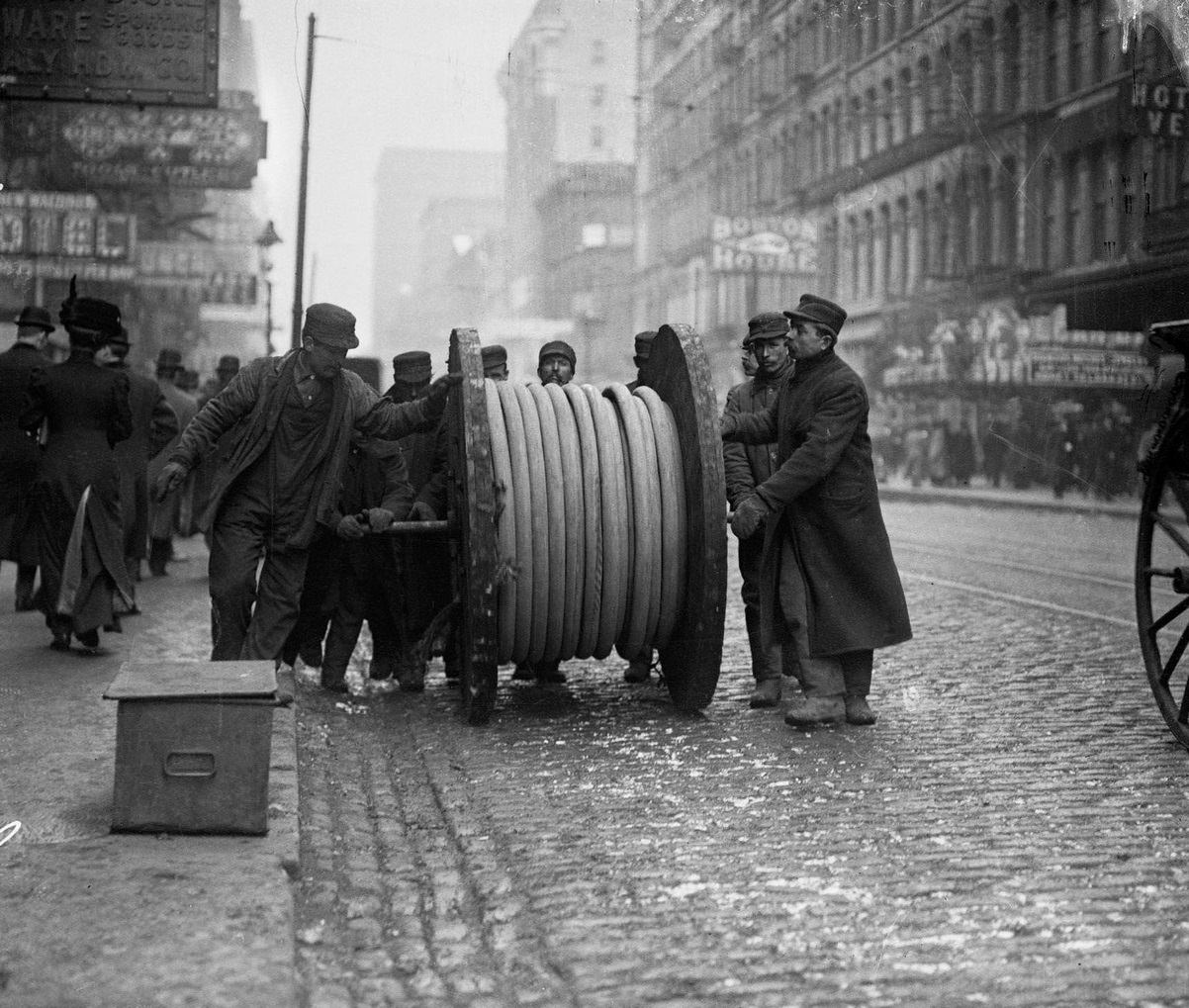 #155 Men rolling a large wheel of underground telephone cable on a business street, Chicago, Illinois, 1910s.