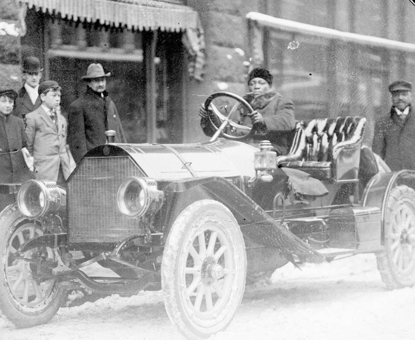 #167 Pugilist Jack Johnson sitting in the driver’s seat of a convertible automobile and holding on to the steering wheel on a snow-covered street in Chicago, Illinois, 1910s.
