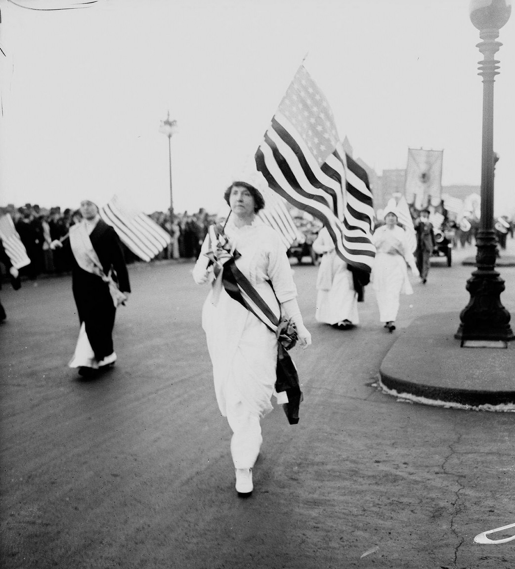 #41 Grace Wilbur Trout wearing a sash, holding an American flag, marching north on South Michigan Avenue, with other suffragists holding flags marching nearby in a women’s suffrage parade in the Loop community area of Chicago, Illinois, 1910s