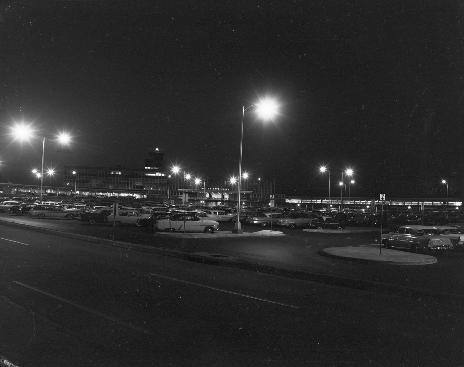 #187 Dallas Love Field at night, 1950. There are lampposts with the lights on. There are automobiles parked on the parking lot.