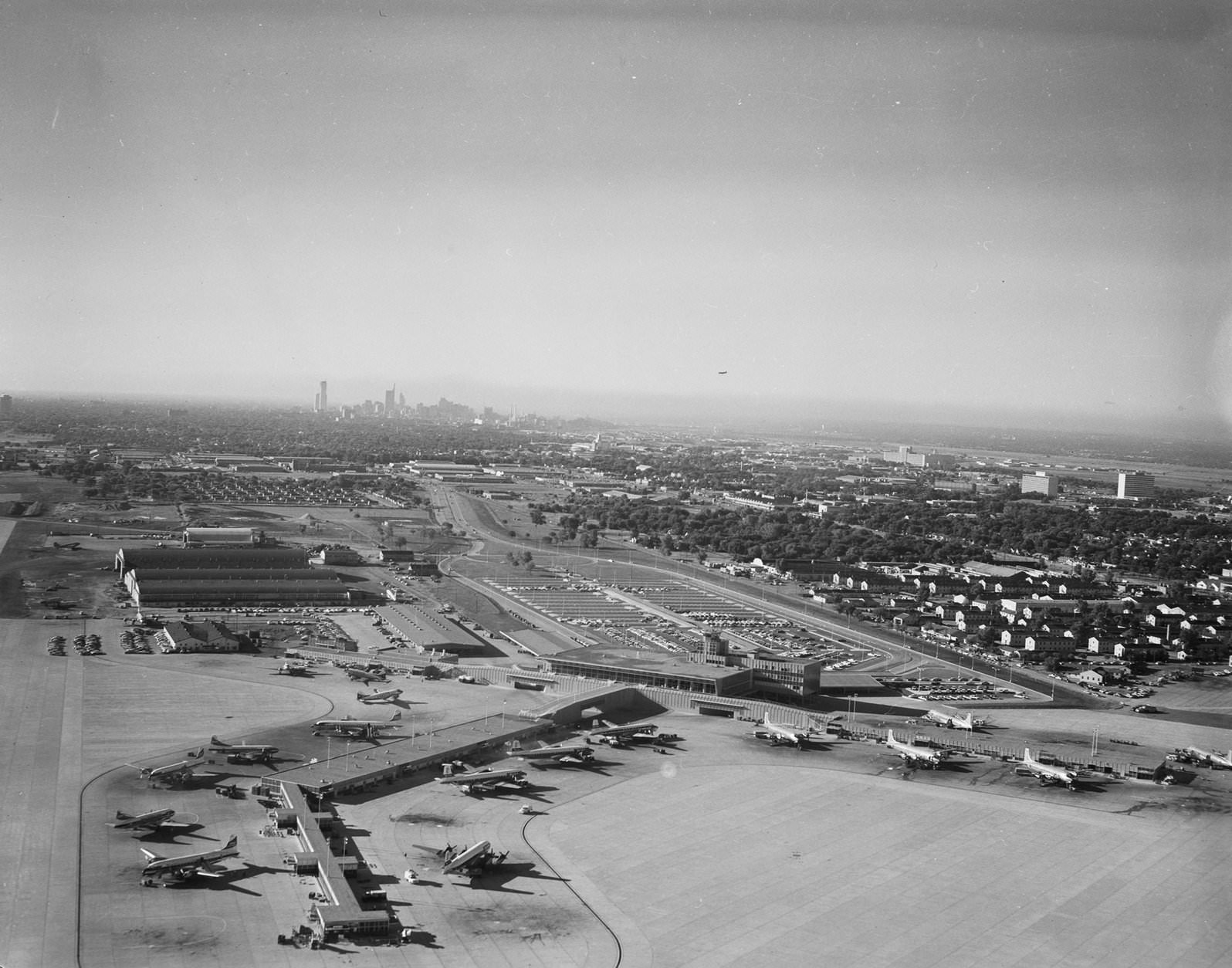 #29 Dallas Love Field,1950. There are airplanes parked on the parking lot. There are a lot of buildings on the background.
