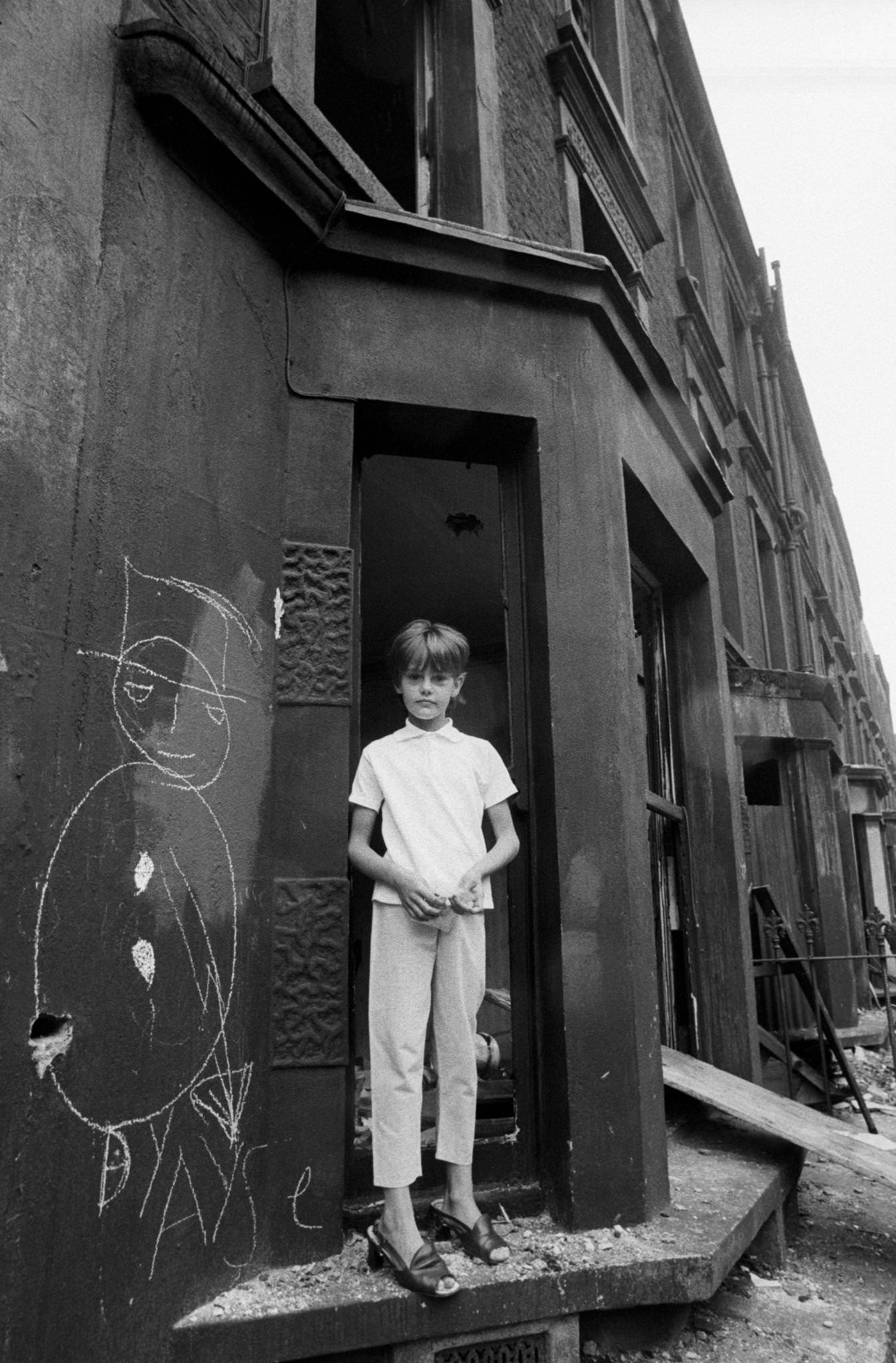 #56 A thin child wearing women’s shoes standing in a doorway next to a graffiti figure in slum housing in the notorious Gorbals district of Glasgow in 1969.